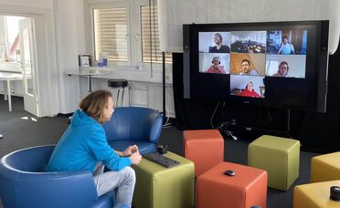 Ein Mitarbeiter sitzt in einem modernen Büro und nimmt an einer Videokonferenz teil, bei der mehrere Kollegen auf einem großen Bildschirm zu sehen sind.