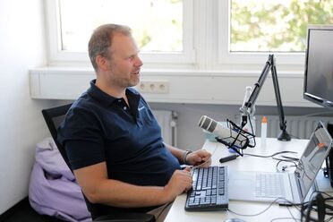 Oliver Schulze sitzt an einem Schreibtisch mit Laptop und Mikrofon, während er an einem Interview teilnimmt.