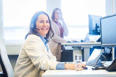 Anne Klein sitzt am Schreibtisch mit Headset und arbeitet an einem Open Source DMS, im Hintergrund ein weiterer Mitarbeiter.