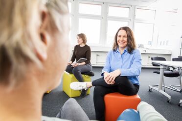 Zwei Geschäftsleute sitzen auf Sitzwürfeln in einem modernen Büro und besprechen sich, während eine dritte Person im Vordergrund zuhört.