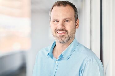 Ein Mitarbeiter steht vor einem Fenster, trägt ein hellblaues Hemd und erklärt die Backup-Sicherung im Dokumentenmanagement.