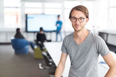 Ein Mitarbeiter steht im Büro vor einem großen Bildschirm, der eine Präsentation zeigt, während Kollegen im Hintergrund arbeiten.
