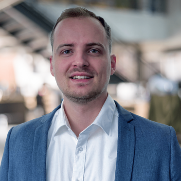 Timm Soworka, Consultant bei GODYO, trägt einen blauen Anzug und ein weißes Hemd, steht in einem modernen Gebäude.
