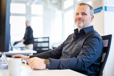 Ein Mitarbeiter sitzt an einem Schreibtisch im Büro und arbeitet am Computer, im Hintergrund ist ein weiterer Mitarbeiter zu sehen.