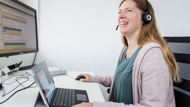 Eine Mitarbeiterin namens Daniela Lutz arbeitet an ihrem Schreibtisch mit einem Laptop und einem Monitor, die das Dokumentenmanagement-System anzeigen.