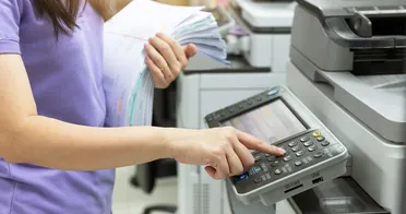 Eine Frau bedient einen Drucker mit einem Stapel Papier in der Hand. Der Workflow wird digitalisiert, um Medienbrüche zu vermeiden.