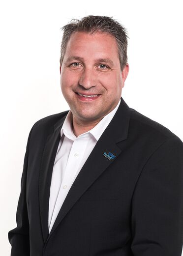 Heinz Kaufmann trägt einen schwarzen Anzug mit einem SelectLine-Pin, steht vor weißem Hintergrund.