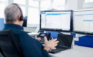 Ein Mitarbeiter mit Headset sitzt an einem Schreibtisch und arbeitet an einem Dokumentenmanagement-System (DMS) mit zwei Monitoren und einem Laptop.