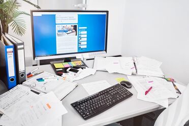 Ein Büroarbeitsplatz zeigt einen Computerbildschirm mit Dokumentenmanagement-Software und viele physische Dokumente auf dem Schreibtisch.