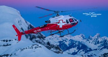 Ein Hubschrauber der Swiss Helicopter Group fliegt über schneebedeckte Berge bei Sonnenuntergang.