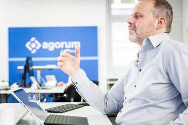 Ein Mitarbeiter zeigt auf ein Dokument auf einem Tablet, im Hintergrund ist das agorum-Logo zu sehen, was IT-Compliance und die Umsetzung von DSGVO und GoBD verdeutlicht.