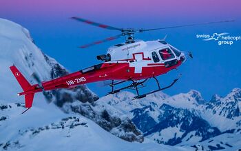 Ein Hubschrauber der Swiss Helicopter Group fliegt über schneebedeckte Berge bei Sonnenuntergang.