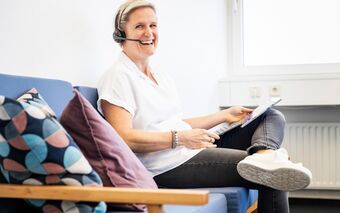 Eine Mitarbeiterin sitzt auf einem blauen Sofa im Büro und arbeitet mit einem Tablet, während sie ein Headset trägt.