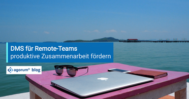 Ein Laptop, eine Sonnenbrille, ein Smartphone und ein Notizbuch liegen auf einem Tisch am Wasser, symbolisieren die Nutzung eines Dokumentenmanagementsystems für Remote-Teams.